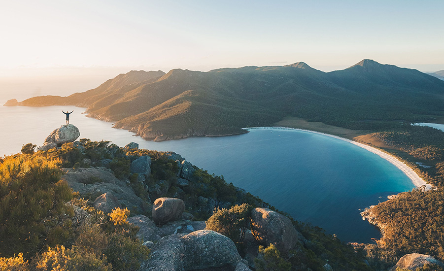 A person stands atop a boulder on a huge cliff, dropping down below to reveal a beautiful ocean bay with a mountainous background.