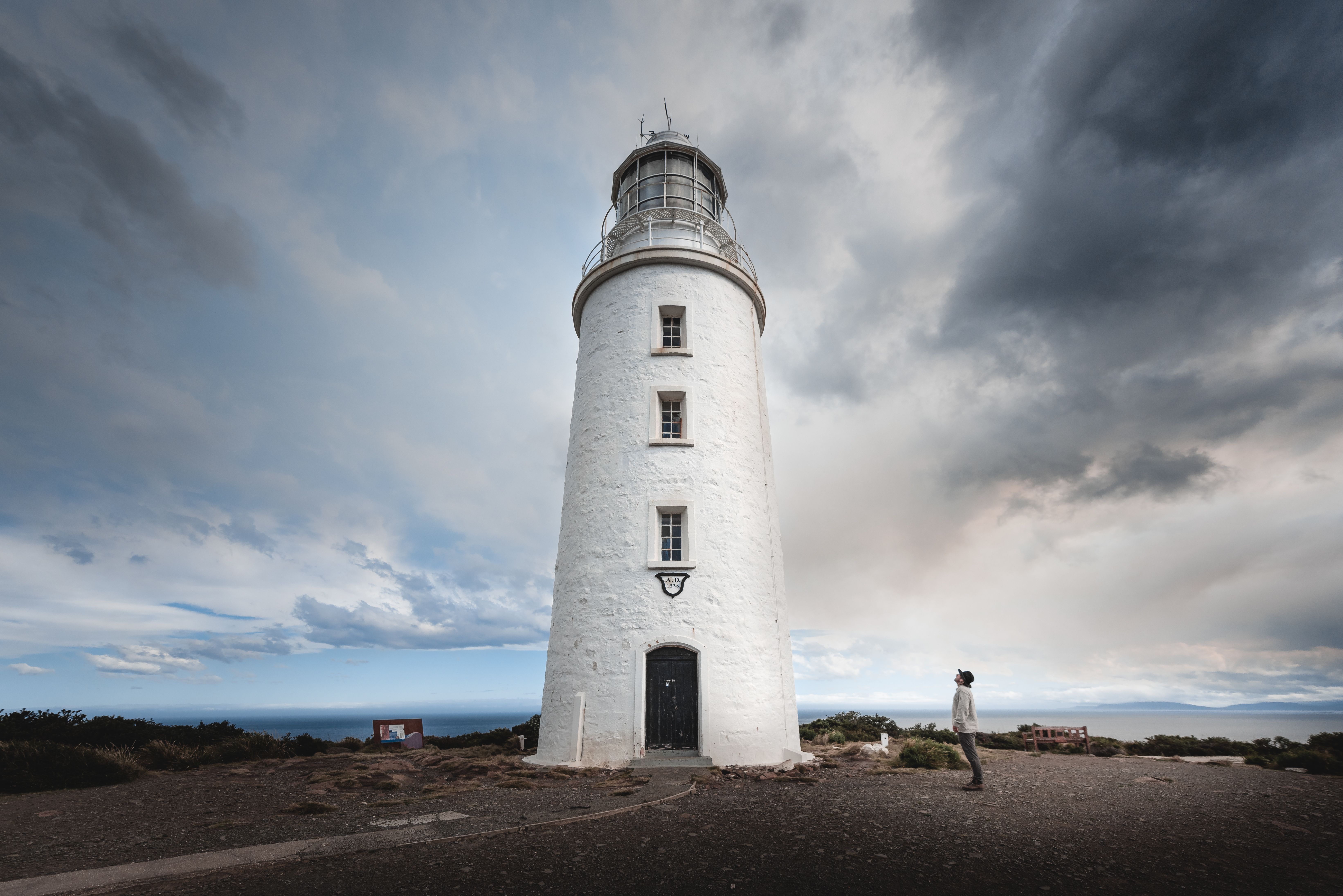 Person looking up at the historic white Cape Bruny Lighthouse under a cloudy sky on Bruny Island
